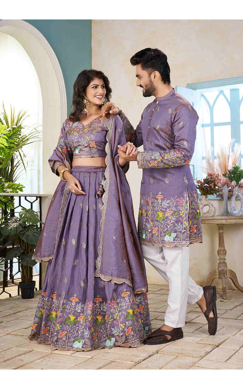 Couple in matching purple traditional outfits standing in a decorated room.