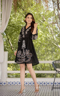 Woman in a black dress with white embroidery standing outdoors.