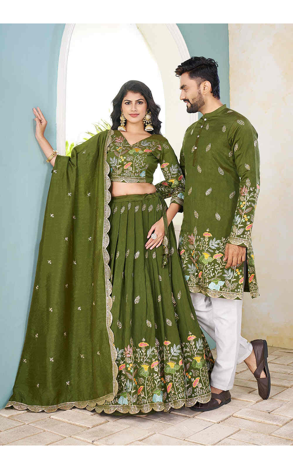 A Couple in mehendi green traditional outfits standing against a light blue wall.