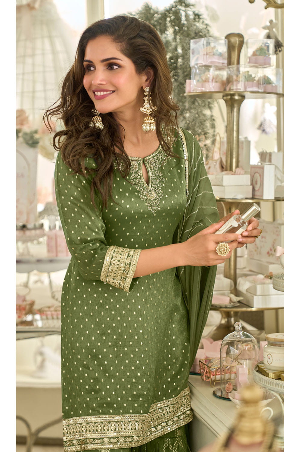 Woman in a green traditional outfit with white patterns, holding a phone, in a decorated indoor setting.