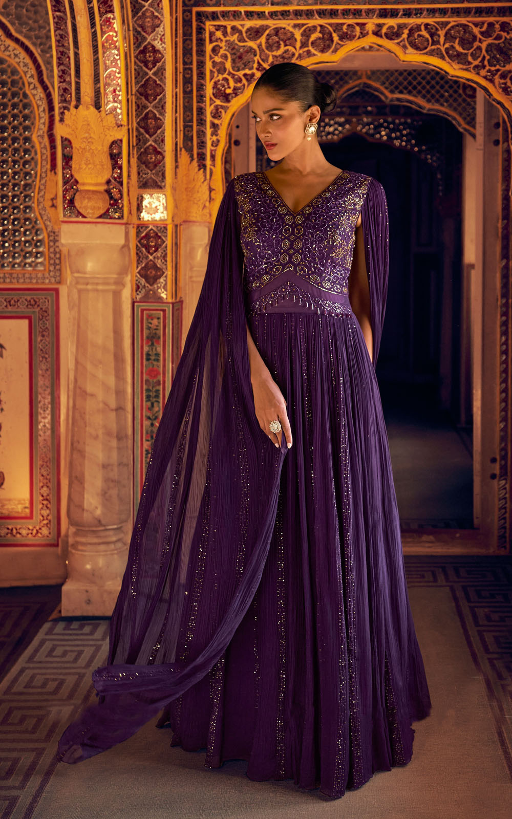 Woman in a purple embroidered gown standing in an ornate room.
