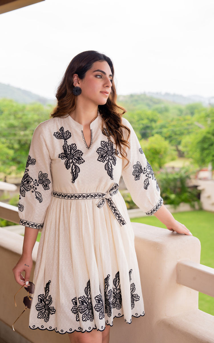 Woman wearing a white dress with black floral embroidery in an outdoor setting.