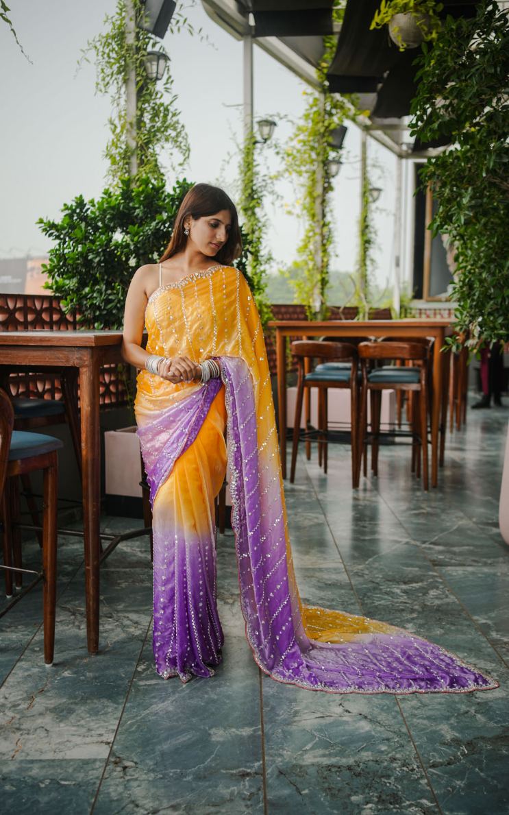 Woman in a yellow and purple saree standing in an outdoor setting with tables and chairs.