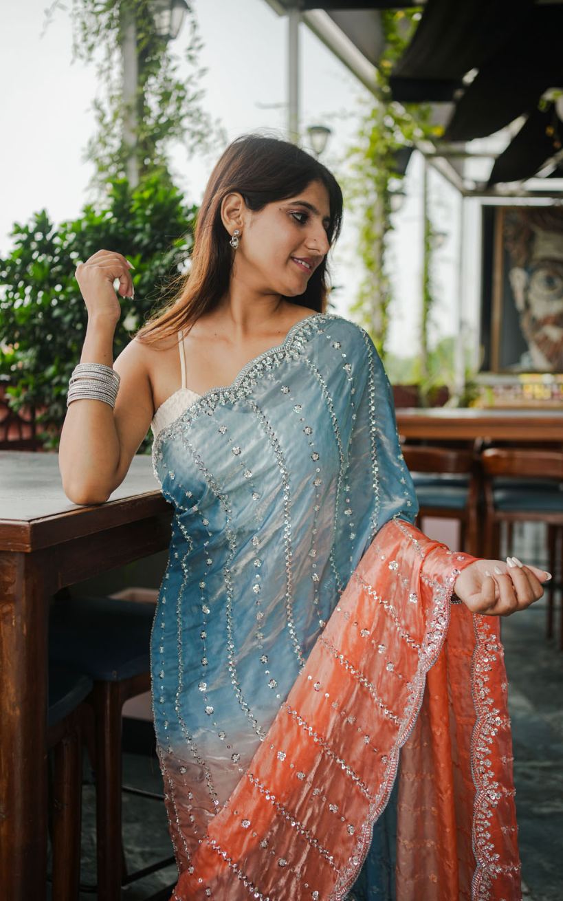 Woman in a light blue saree with a red border standing outdoors.