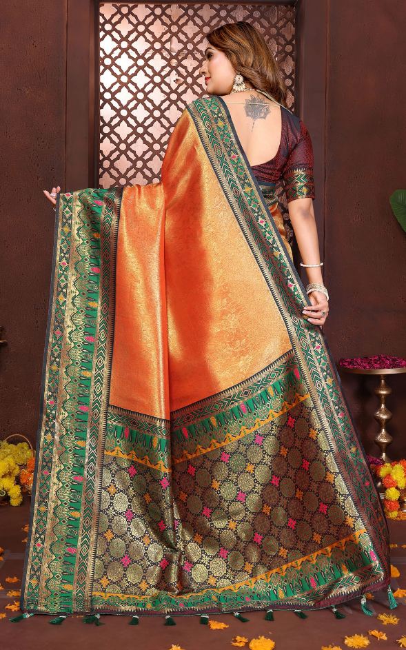 Woman holding an orange saree with green and brown patterns in a decorative setting.