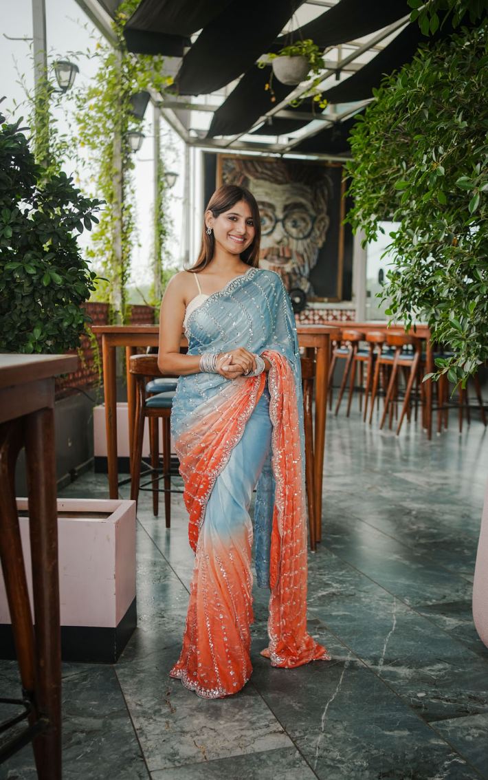 Woman in a light blue and orange saree standing in an outdoor setting with tables and plants.