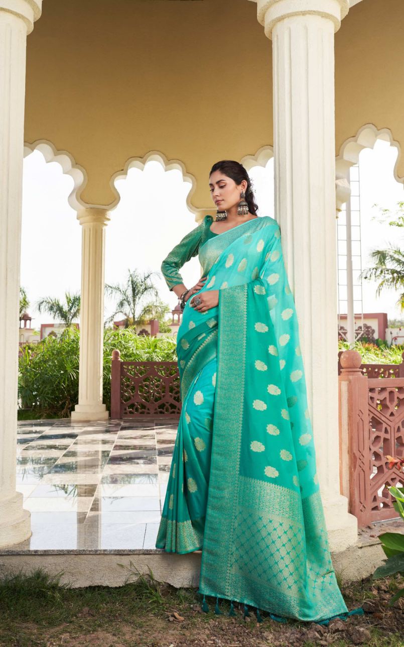 Woman in a teal saree with white patterns standing in an outdoor setting with columns and greenery.