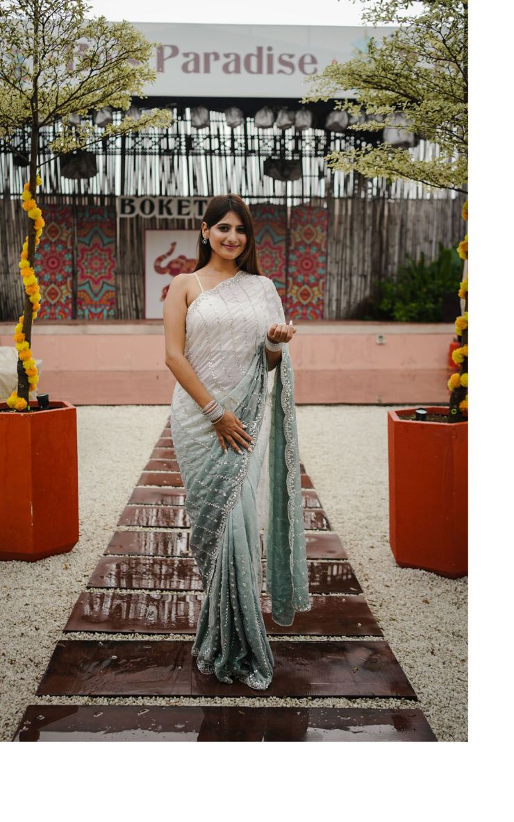 Woman in a saree standing on a decorative path with plants and a 'Paradise' sign in the background.
