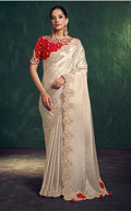 Woman wearing a white saree with red and gold embroidery against a dark green background