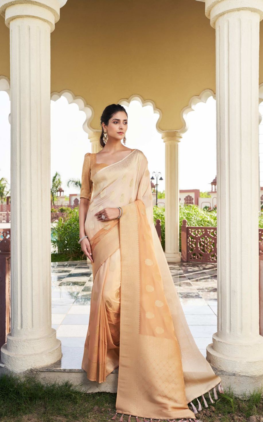 Woman in a beige saree standing in front of classical architecture with columns.