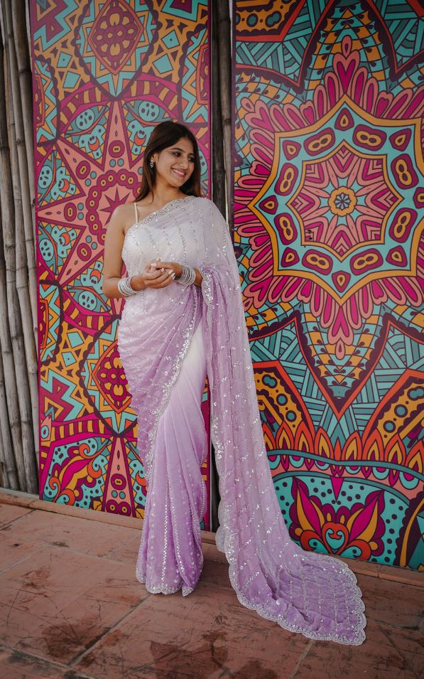 Woman in a white saree standing in front of a colorful geometric patterned wall.