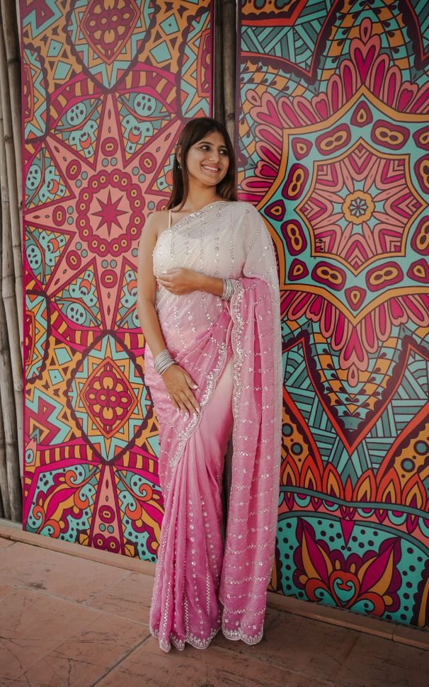 Woman in a pink saree standing in front of a colorful geometric-patterned wall.
