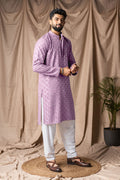 Man wearing a purple kurta with white pants standing against a beige curtain.