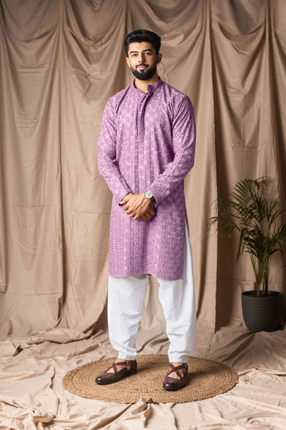 Man wearing a purple kurta with white pants standing against a beige curtain backdrop.