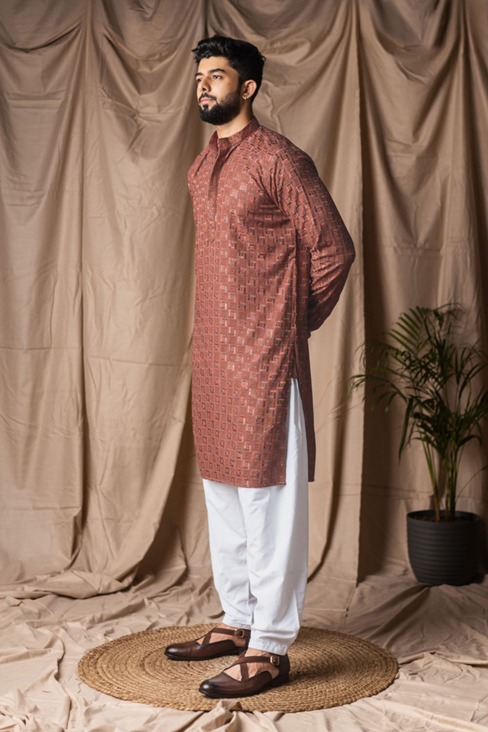 Man wearing a patterned kurta with white pants standing against a beige curtain backdrop.