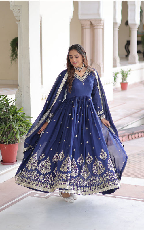 Woman in a blue embroidered traditional outfit standing in a well-lit room with plants.