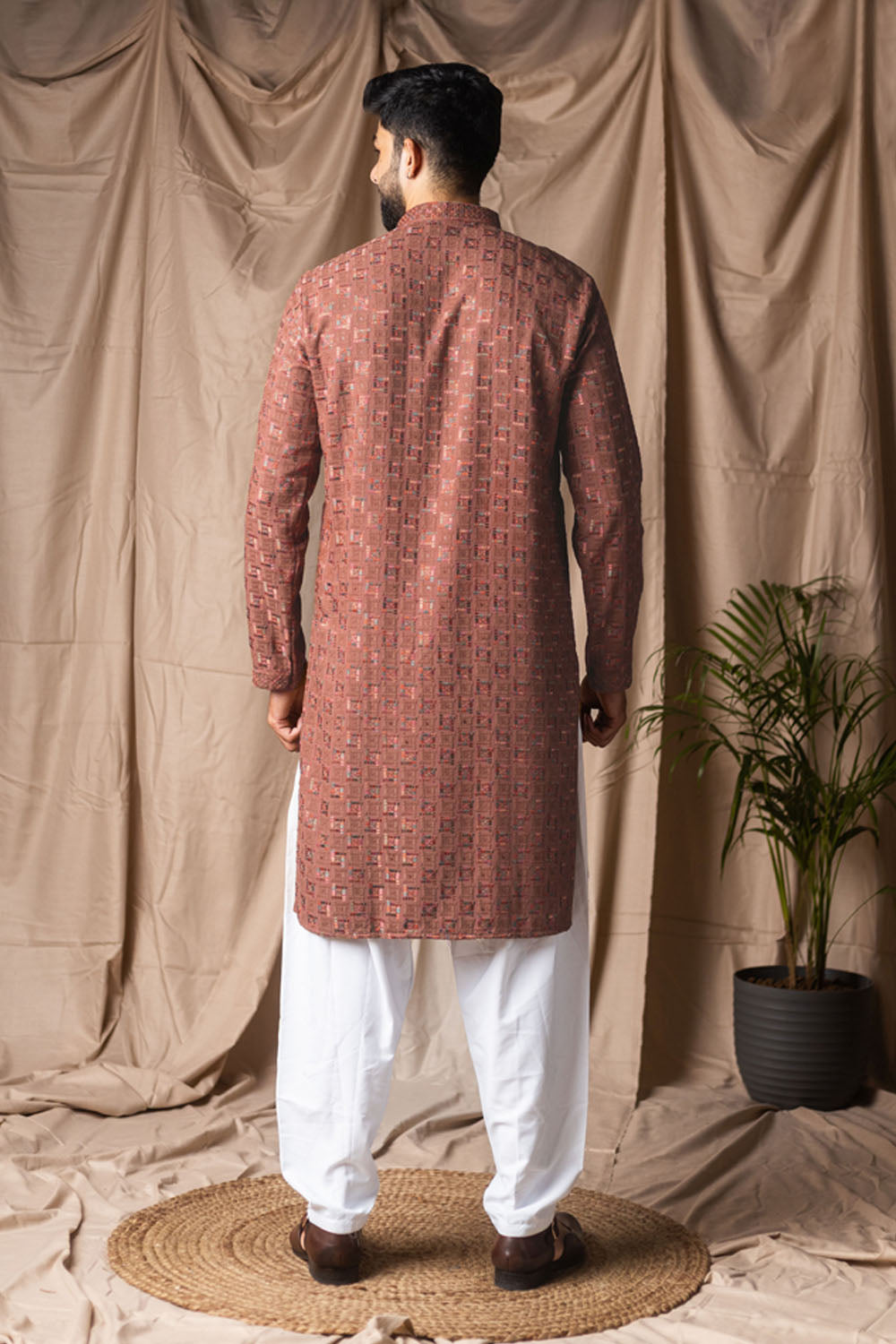 Man wearing a red patterned kurta with white pants standing against a beige curtain backdrop.