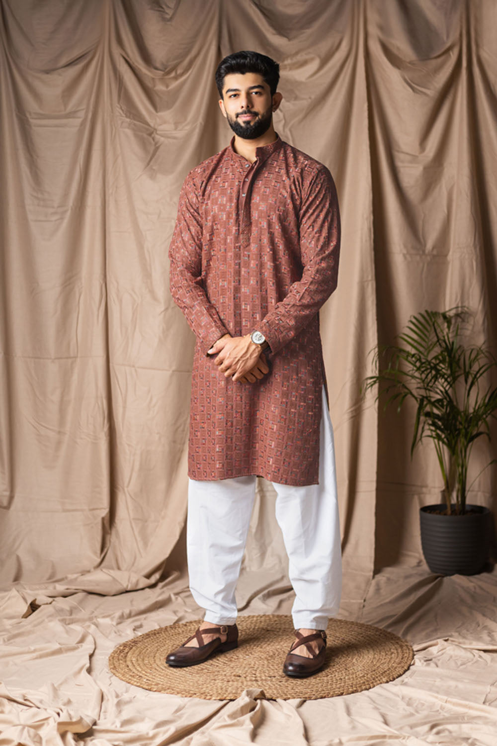 Man wearing a maroon kurta with white pants standing against a beige curtain backdrop.