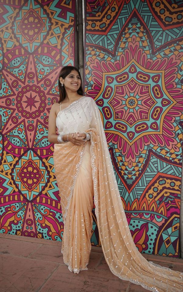 Woman in a beige saree standing against a colorful geometric-patterned wall.