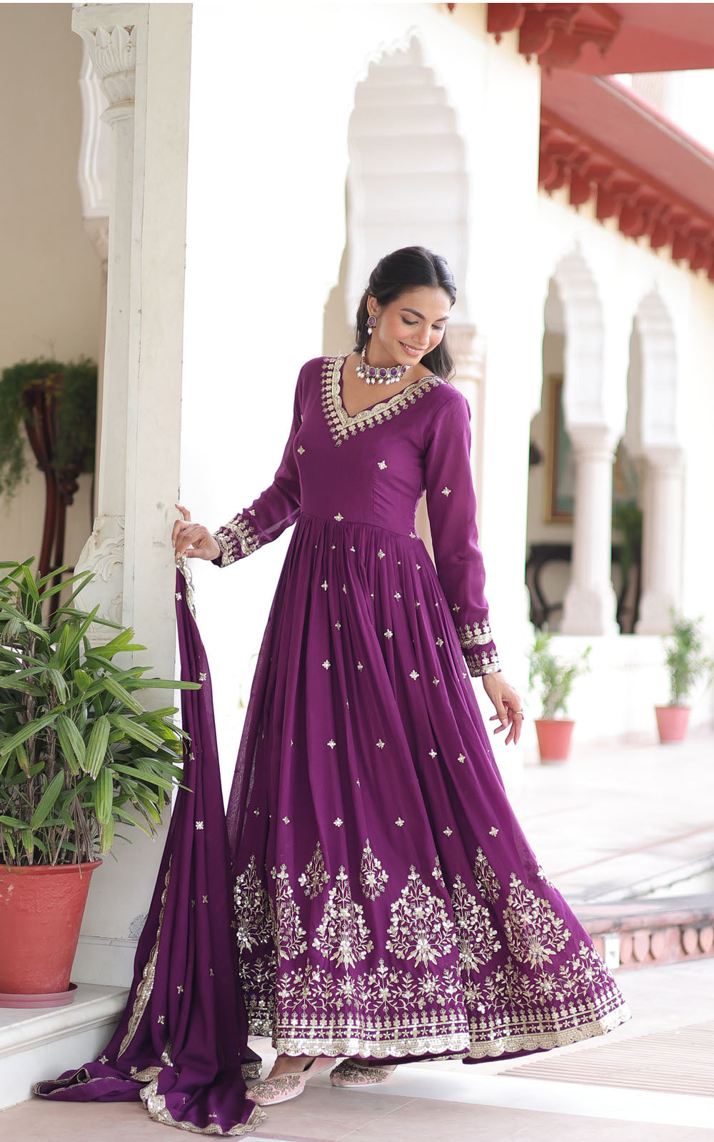 Woman in a purple embroidered anarkali suit standing in a decorative indoor setting.