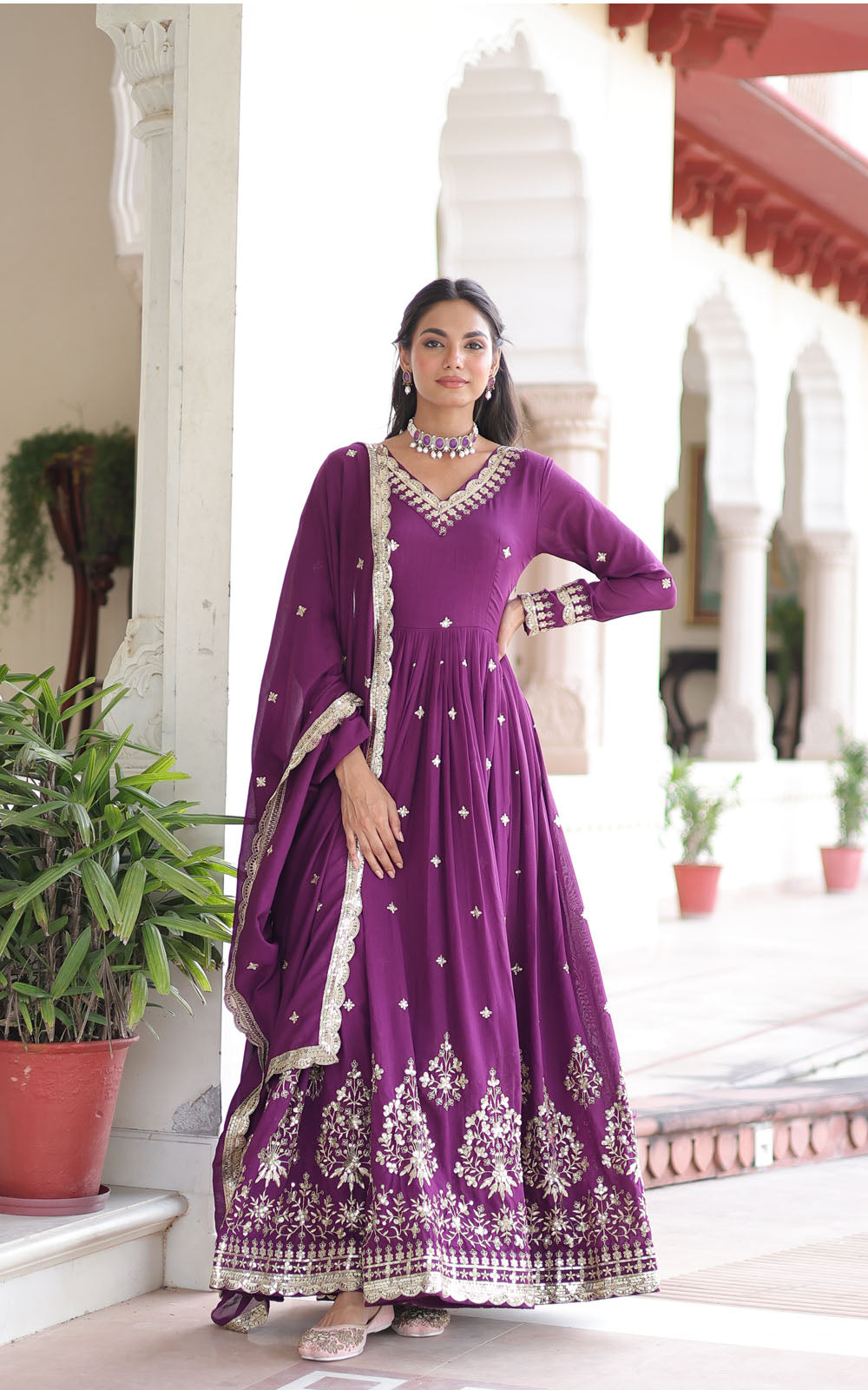 Woman in a purple traditional outfit standing in an elegant indoor setting with plants.