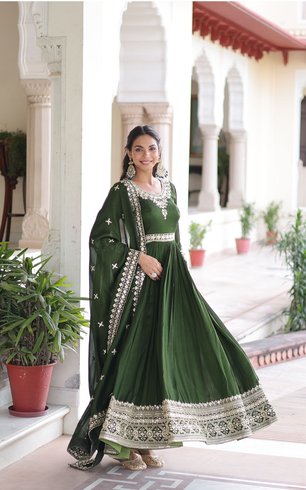 Woman in a green traditional outfit standing in an outdoor setting with plants and architectural elements.