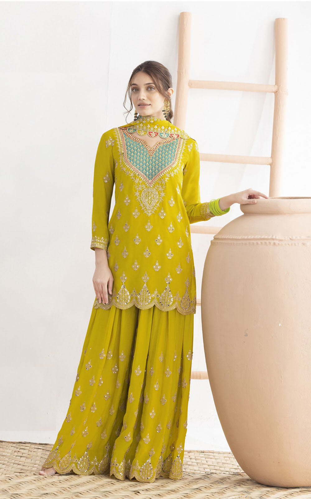 Woman wearing a yellow embroidered traditional outfit standing next to a beige pot on a white background