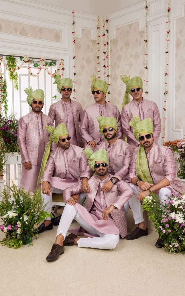 Group of men in pink traditional attire with green turbans in a decorated room.