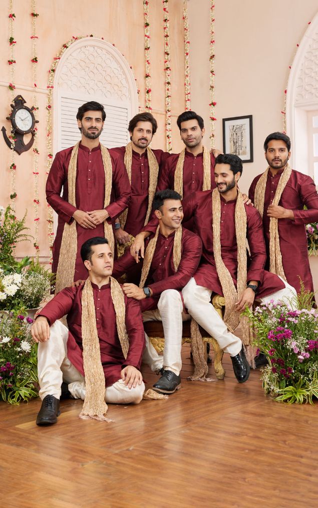 Group of men in maroon traditional attire with long scarves in a decorated indoor setting.