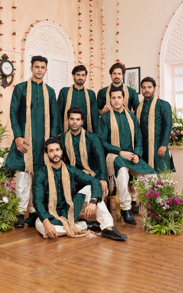 Group of men in green and beige traditional attire posing indoors with decorative elements.