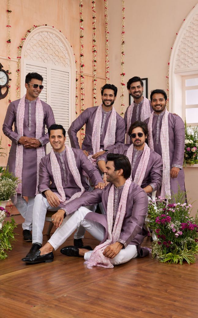 Group of men in matching purple traditional outfits sitting on a wooden floor with decorative elements.