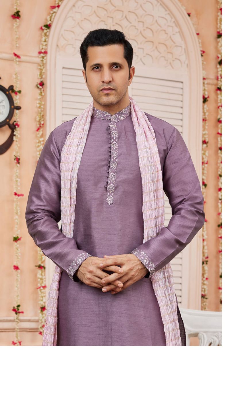 Man wearing a purple traditional outfit with a pink scarf in front of a decorative wall.