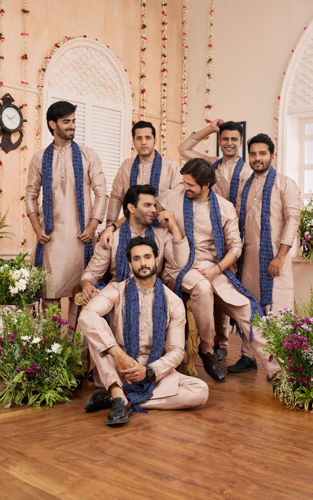 Group of men in traditional attire with blue scarves in a decorated indoor setting.