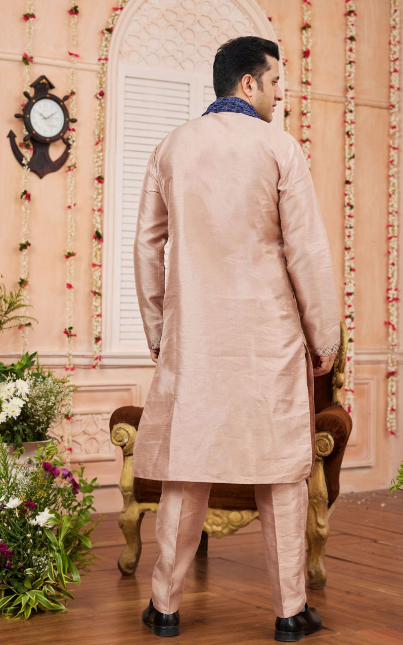 Man wearing a beige sherwani in a decorated room with flowers and a clock.