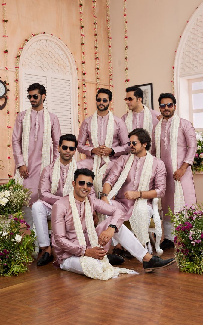 Group of men in pink traditional attire with white scarves in a decorated indoor setting.