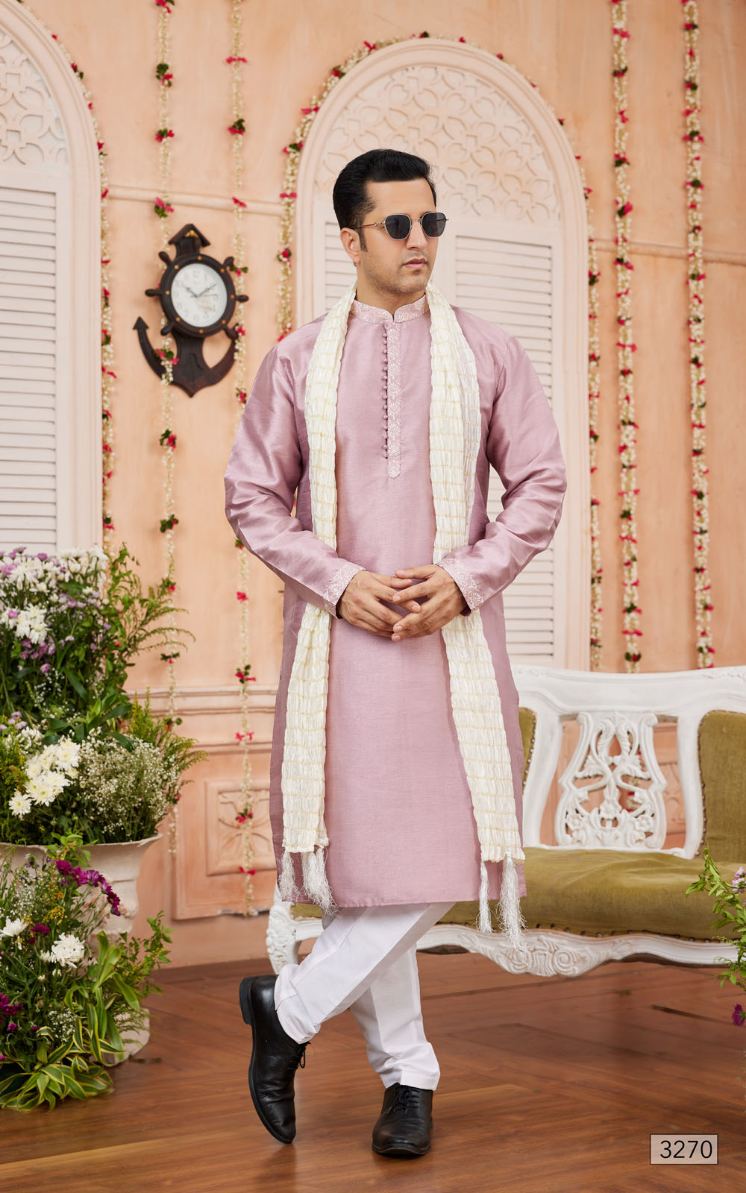 Man in a pink kurta with white pants and scarf standing in a decorated room.