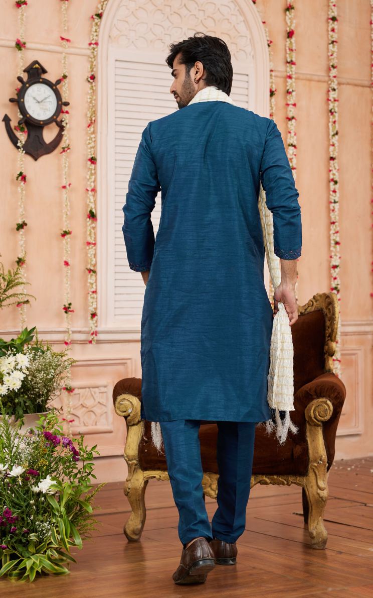 Man in a blue kurta standing in a decorated room with flowers and a clock.
